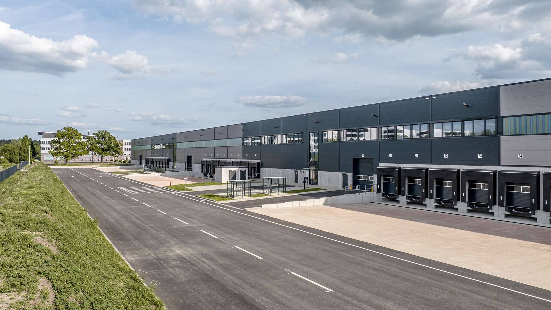 Moderne Lagerhalle mit Laderampen, Parkplatz und angrenzendem Grünbereich unter einem bewölkten Himmel.