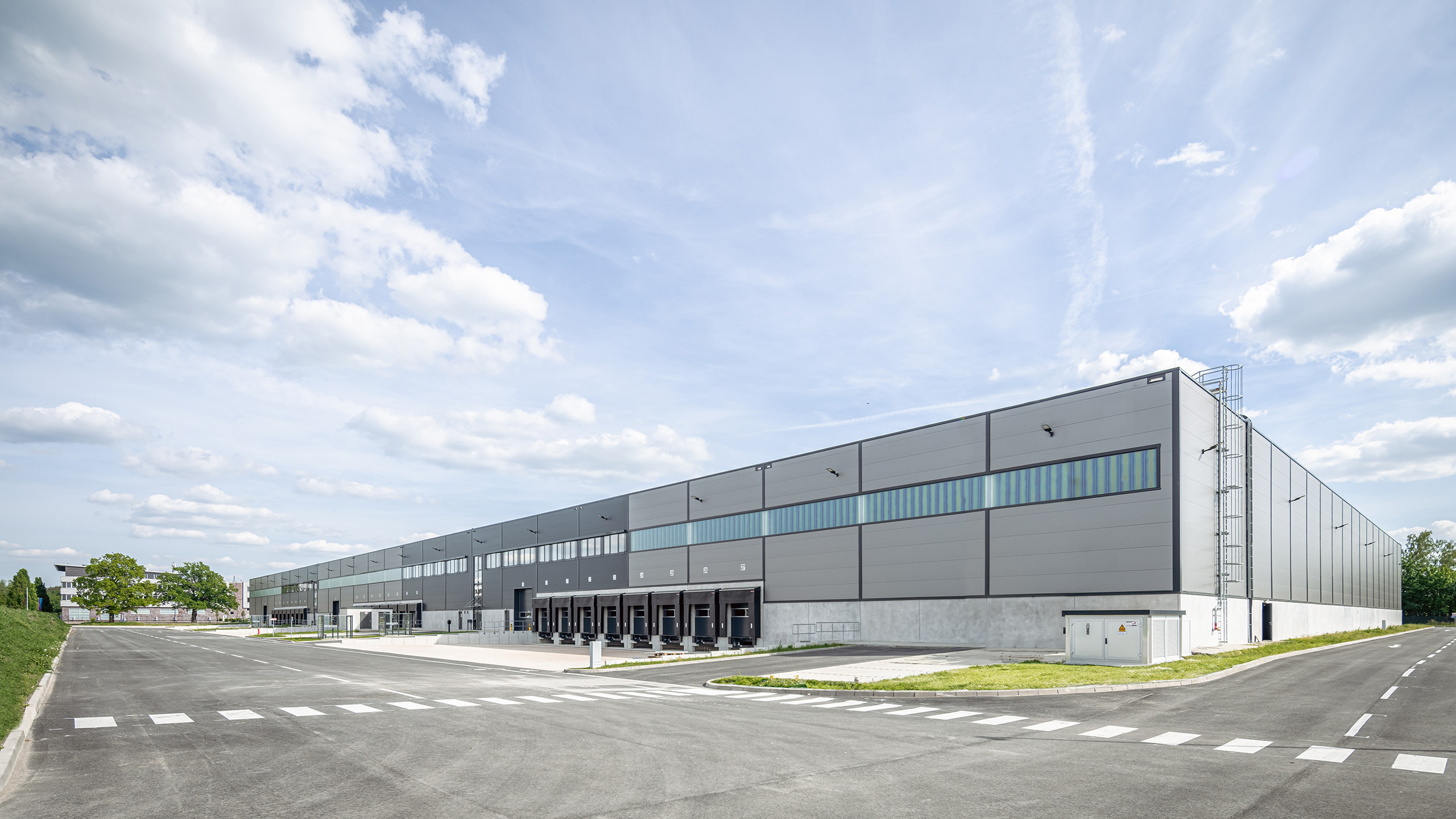 Moderner Lagerhauskomplex mit Lkw-Rampen, grauer Fassade und grünem Umland unter einem bewölkten Himmel.