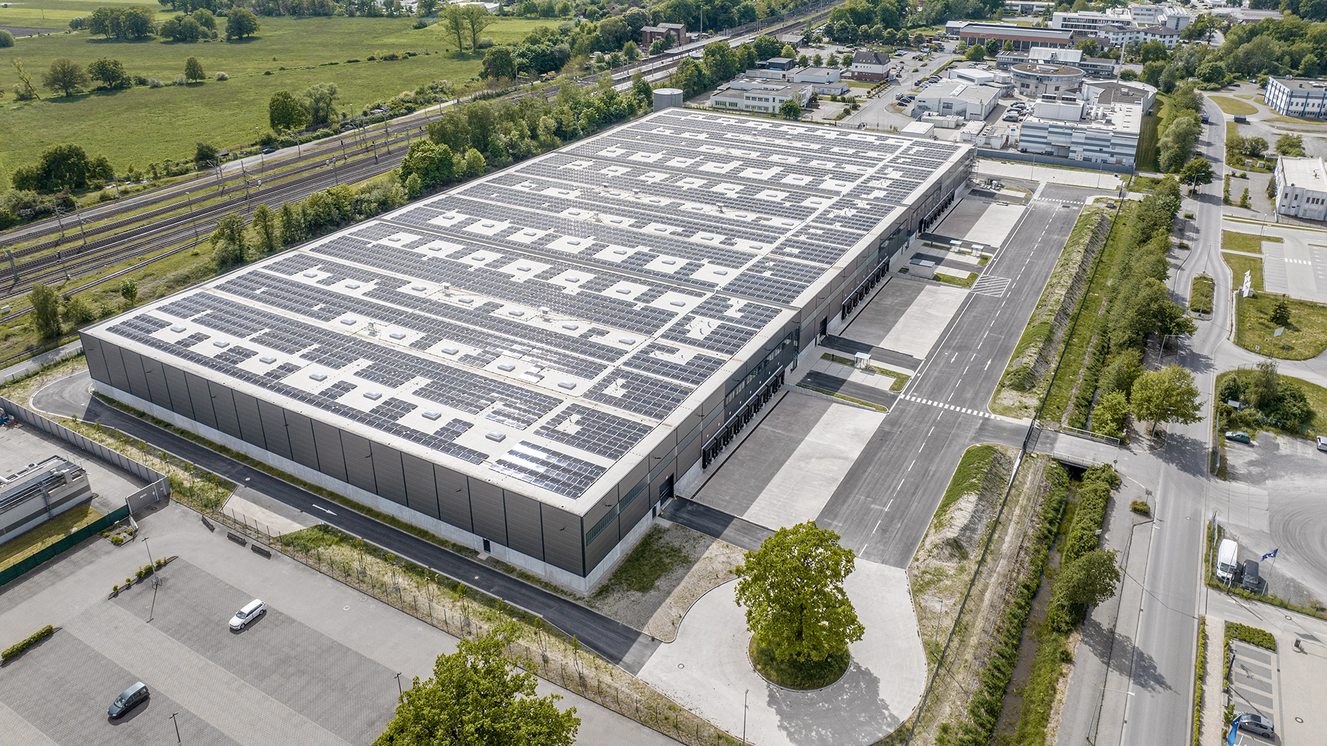 Luftaufnahme eines modernen Logistikgebäudes mit Solarpanelen und umliegenden Parkplätzen und Straßen.