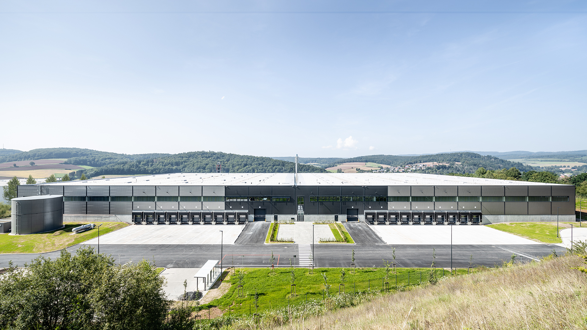 Großes modernes Lagerhaus mit Ladebereichen, umgeben von sanften Hügeln und klarem Himmel.