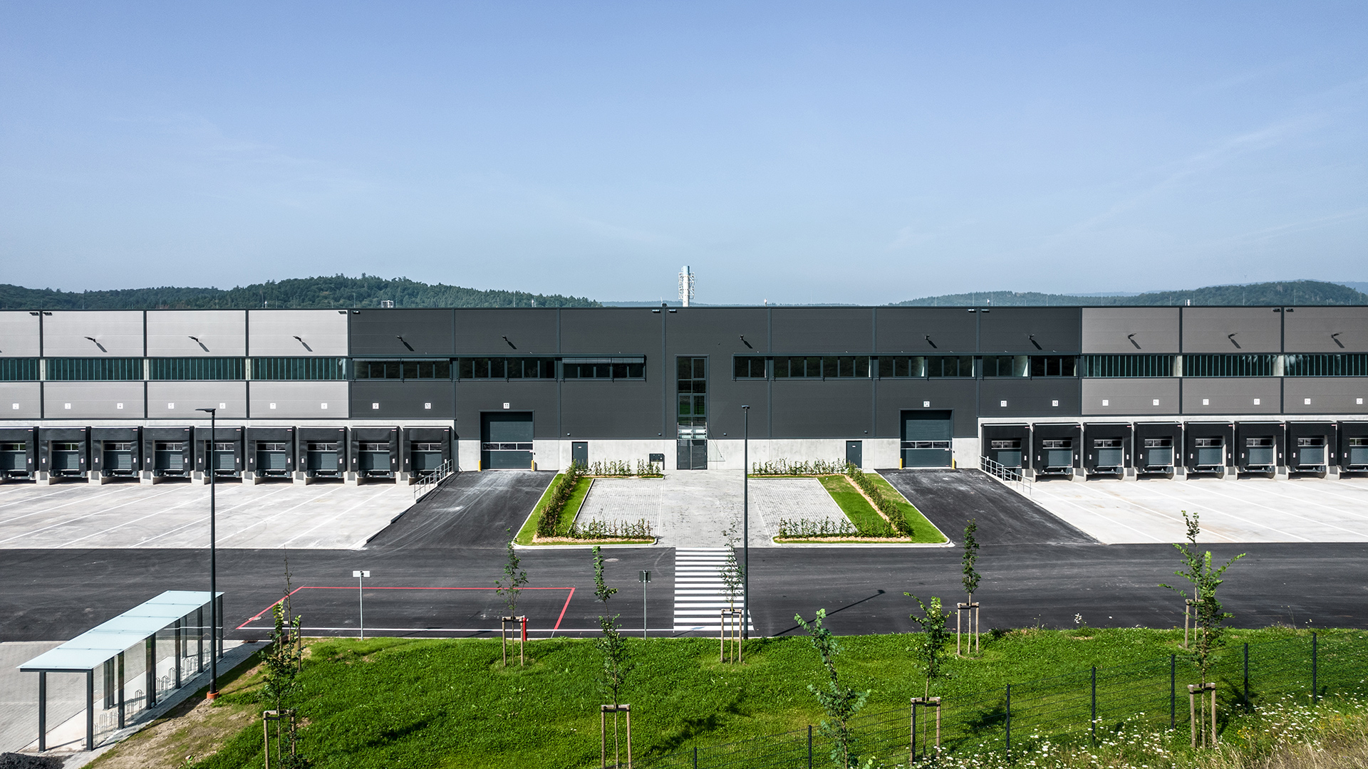 Großes modernes Lagergebäude mit mehreren Laderampen, umgeben von Grünflächen und einem klaren blauen Himmel.