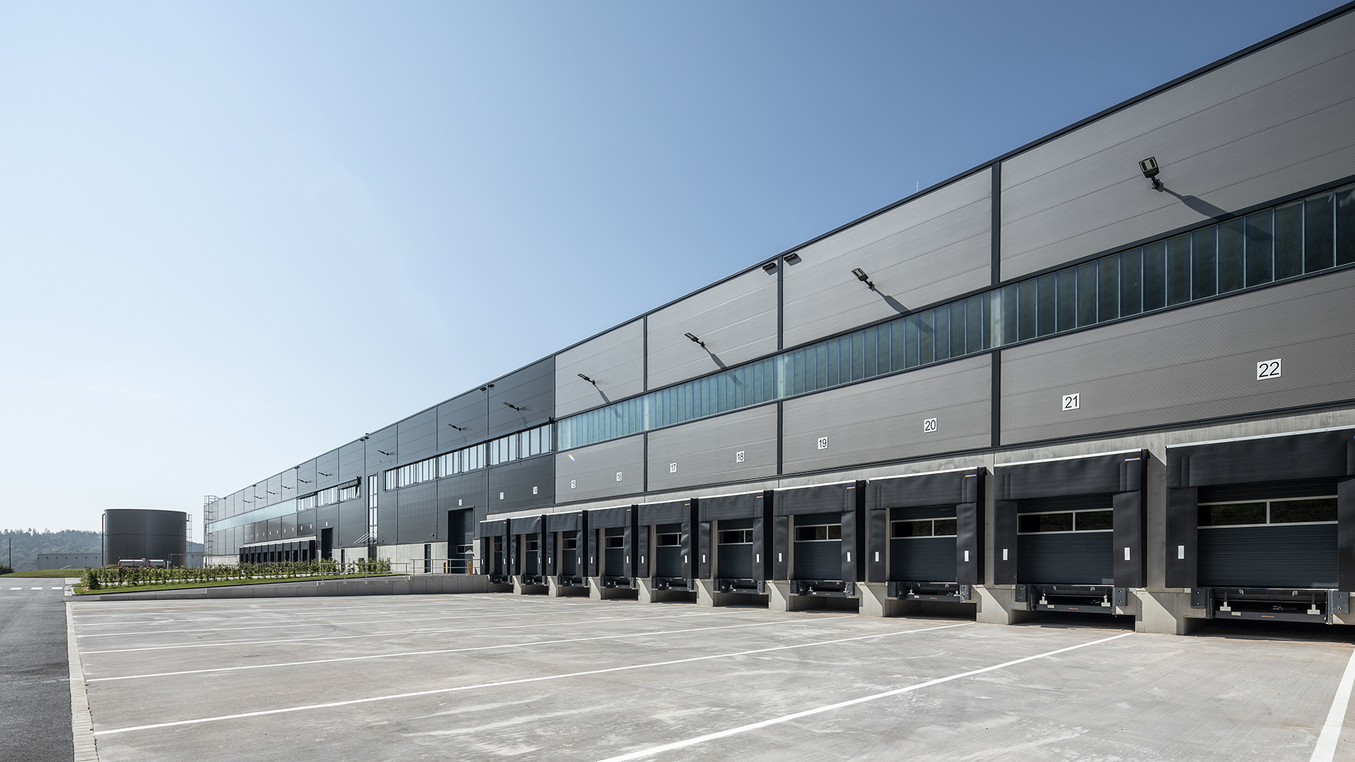 Moderne Lagerhalle mit Verladeplätzen, umgeben von Parkplatz und klar blauem Himmel.