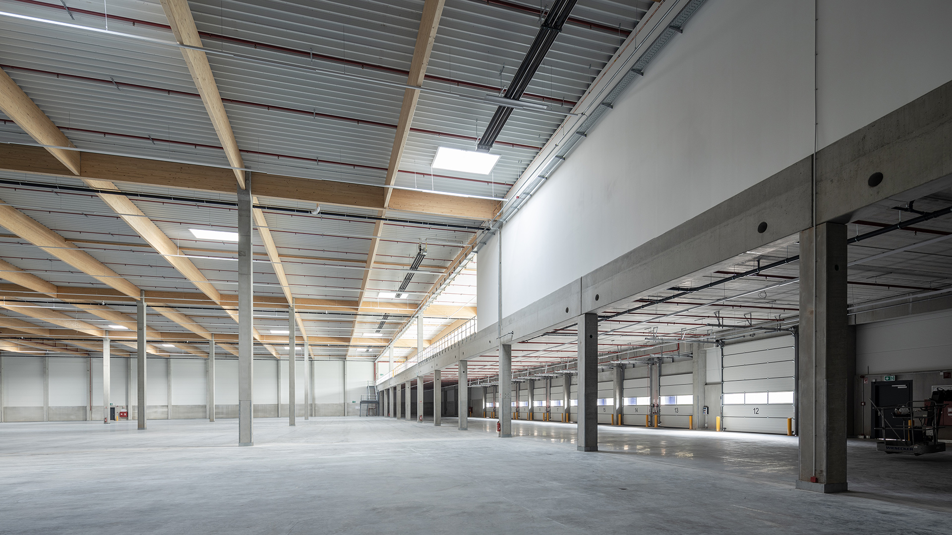 Großer, heller Lagerraum mit Holzbalkendecke und großen Fenstern, grauem Boden und Betonstützen.