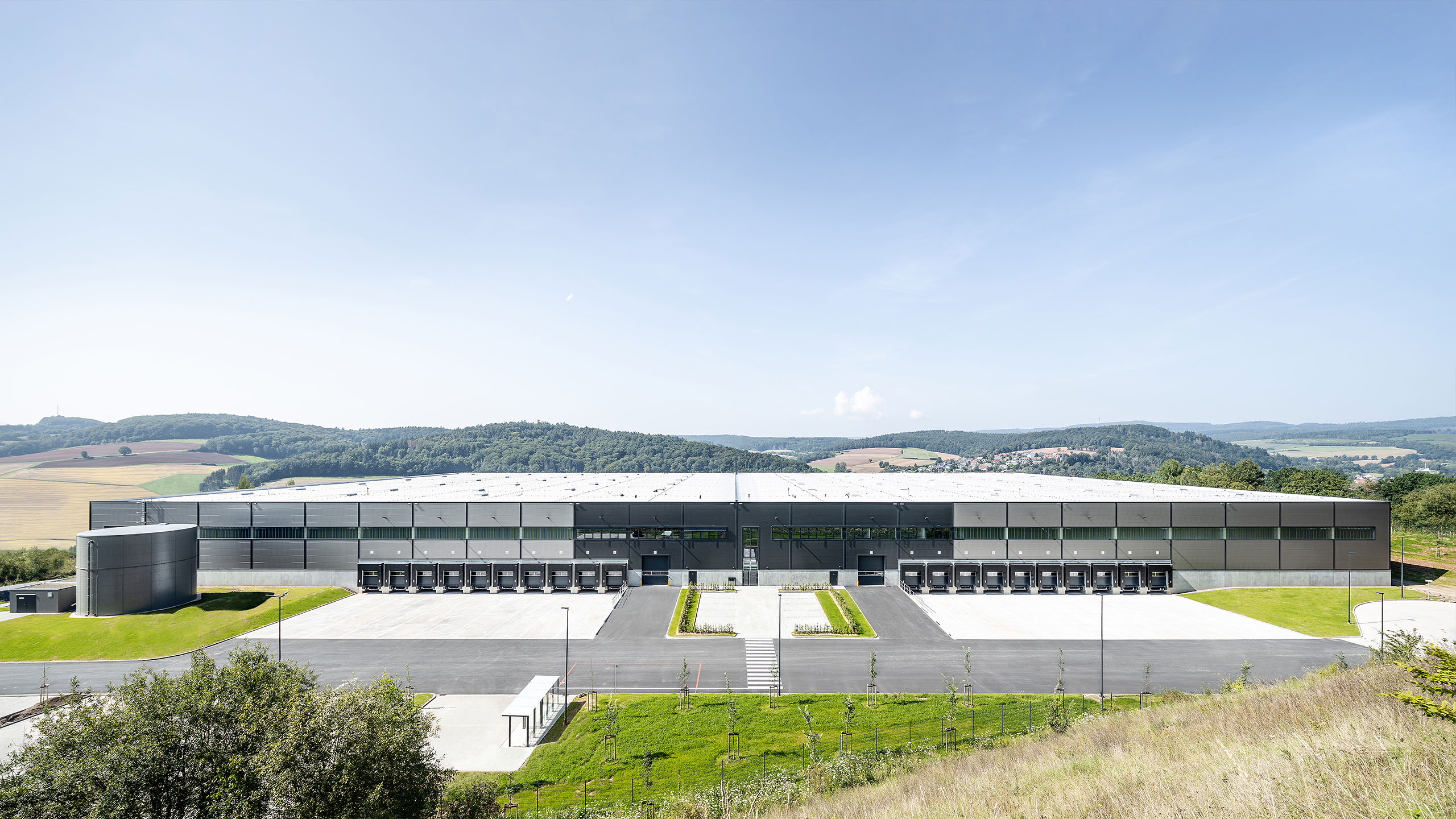 Ein modernes Logistikgebäude umgeben von einer grünen Landschaft unter einem klaren Himmel.