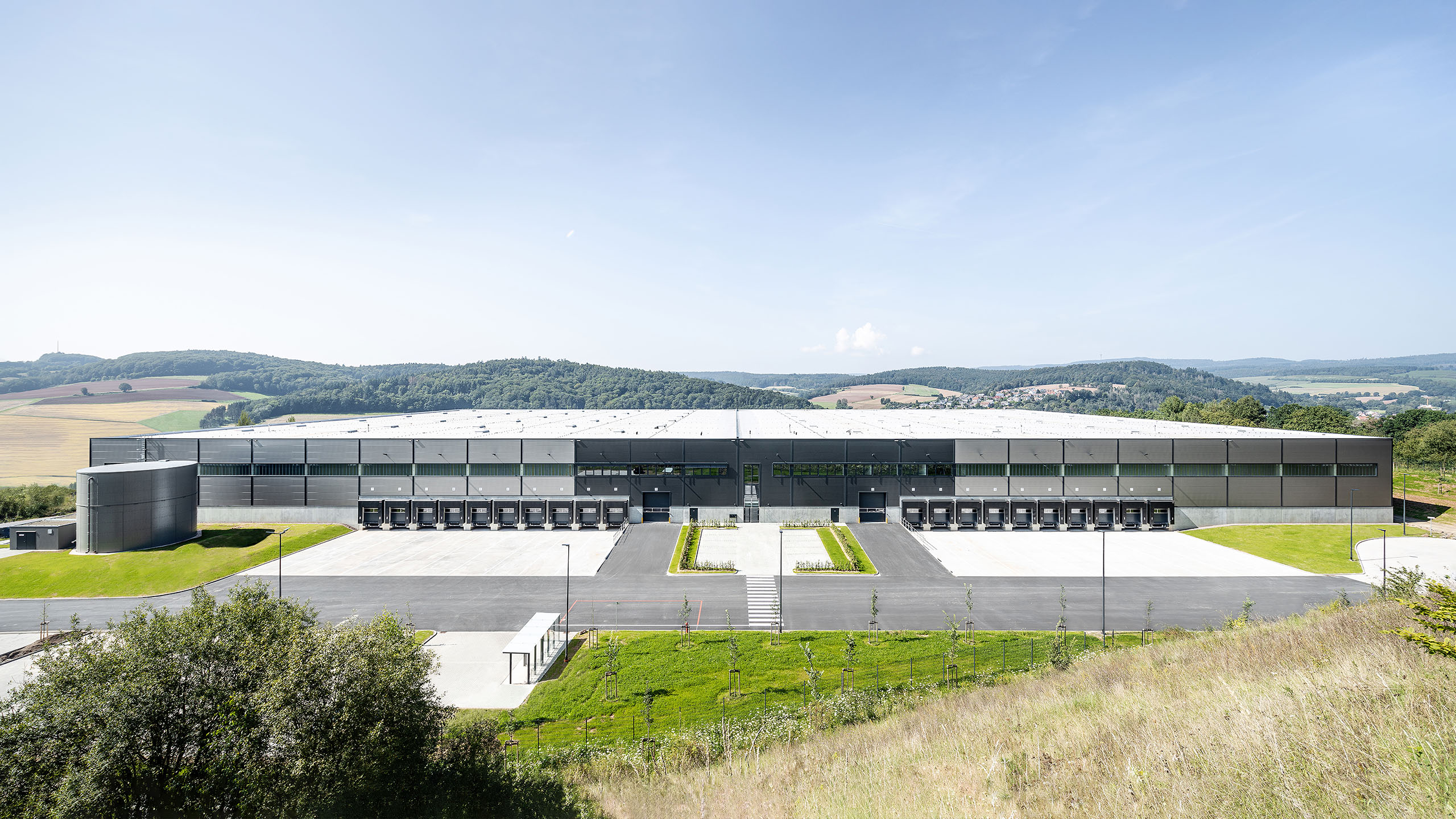 Moderne Lagerhalle mit Ladebuchten, umgeben von grüner Landschaft unter blauem Himmel.