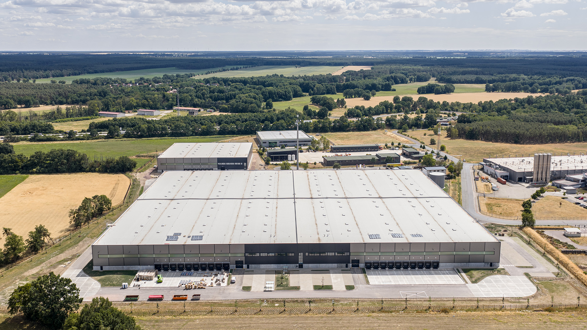 Luftaufnahme eines großen Lagerhauses mit angrenzenden Grünflächen und umliegenden Gebäuden.