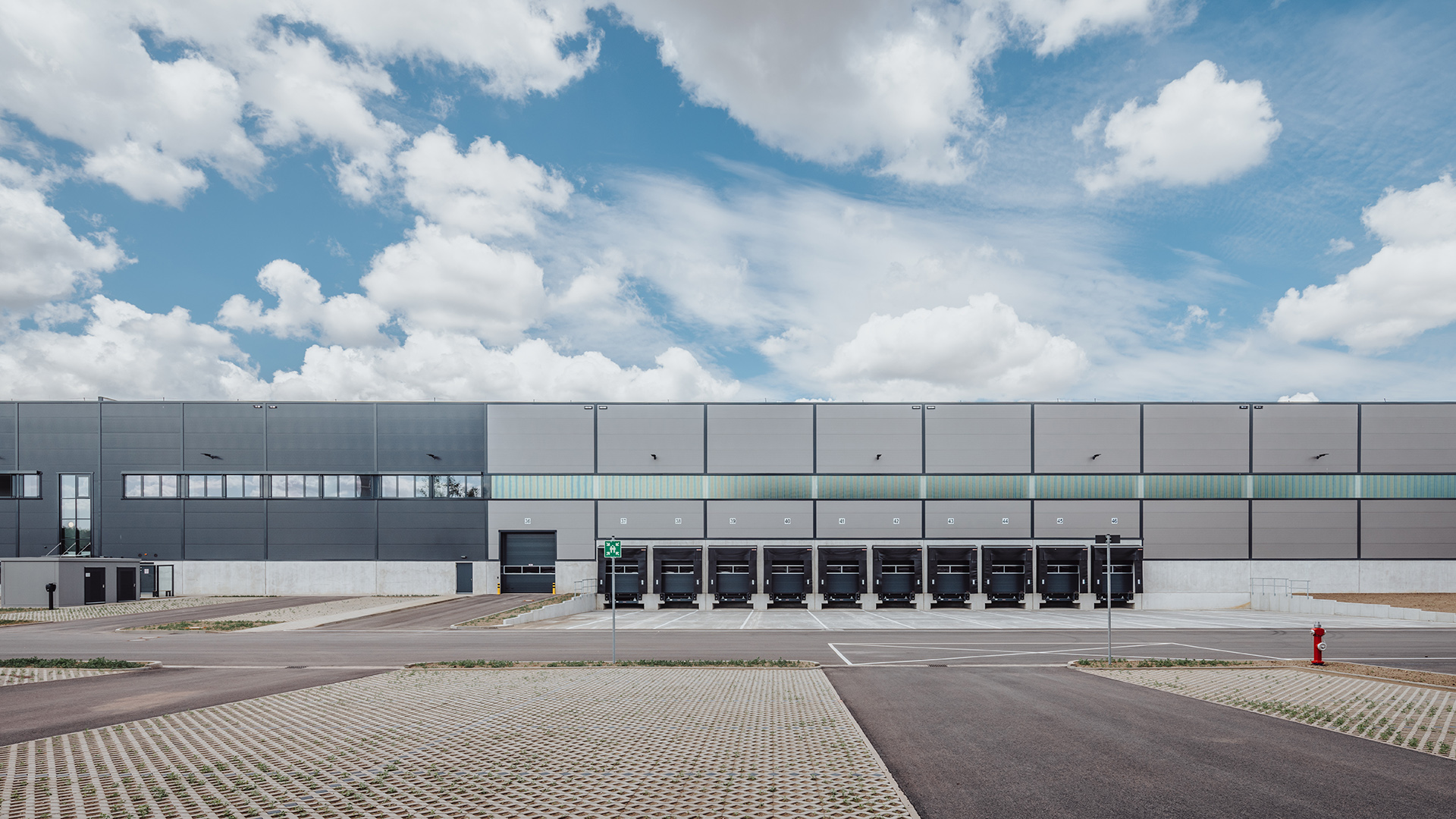 Moderne Lagerhalle mit Ladebucht, Parkfläche und bewölktem Himmel bei Tageslicht.