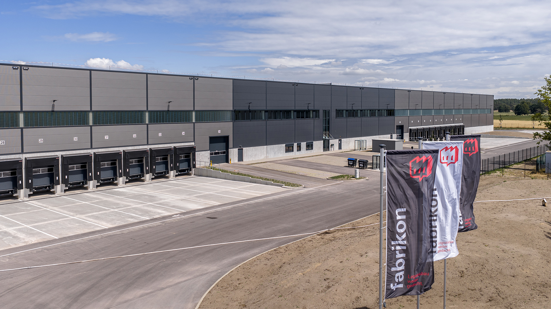 Moderne Lagerhalle mit Ladebucht, geparkten Lkw und Firmenflaggen vor einem blauen Himmel.