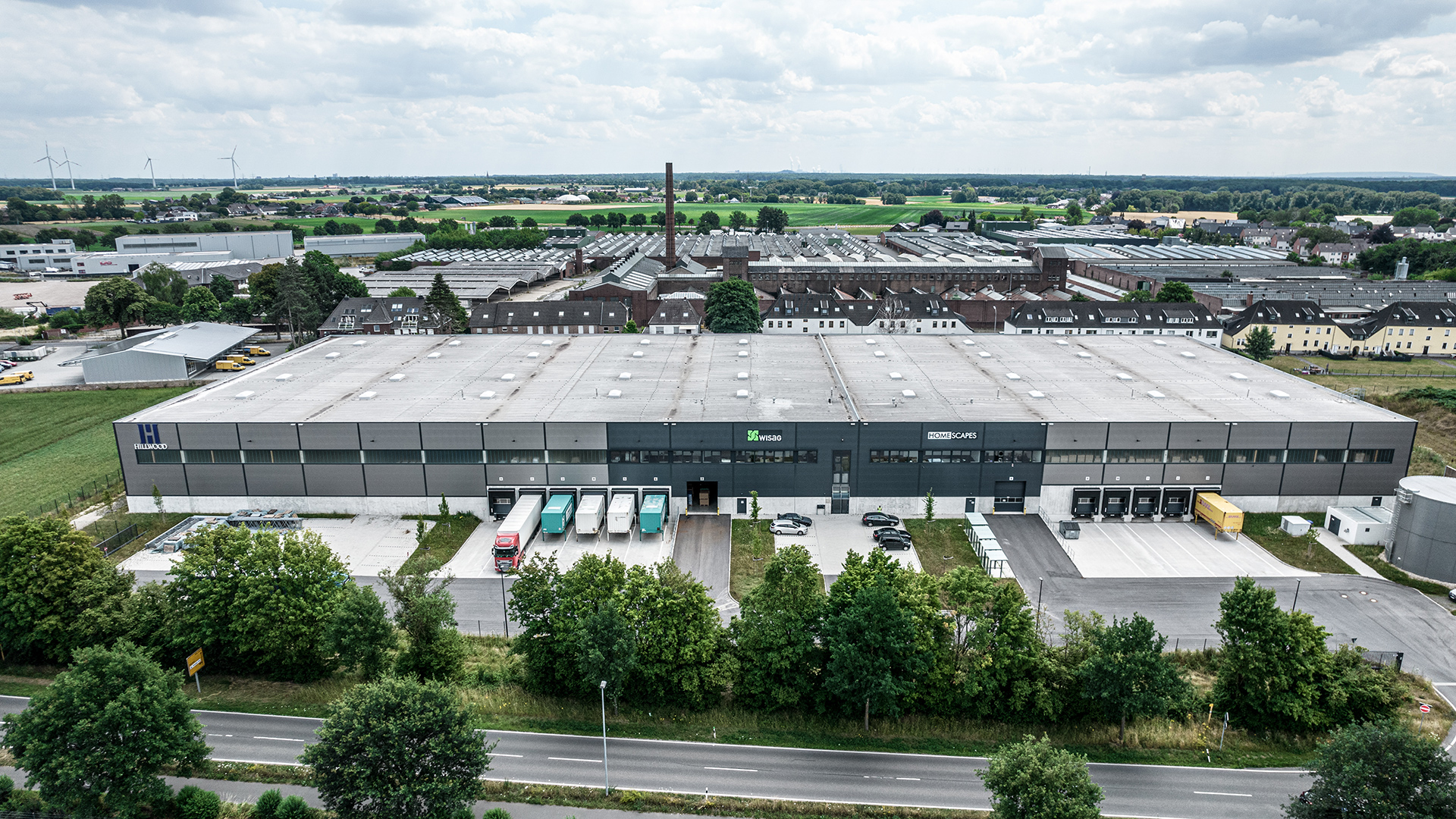 Luftaufnahme eines modernen Lagerhauses mit Lkw, Parkplätzen und umgebender Landschaft, umgeben von Bäumen und Feldern.
