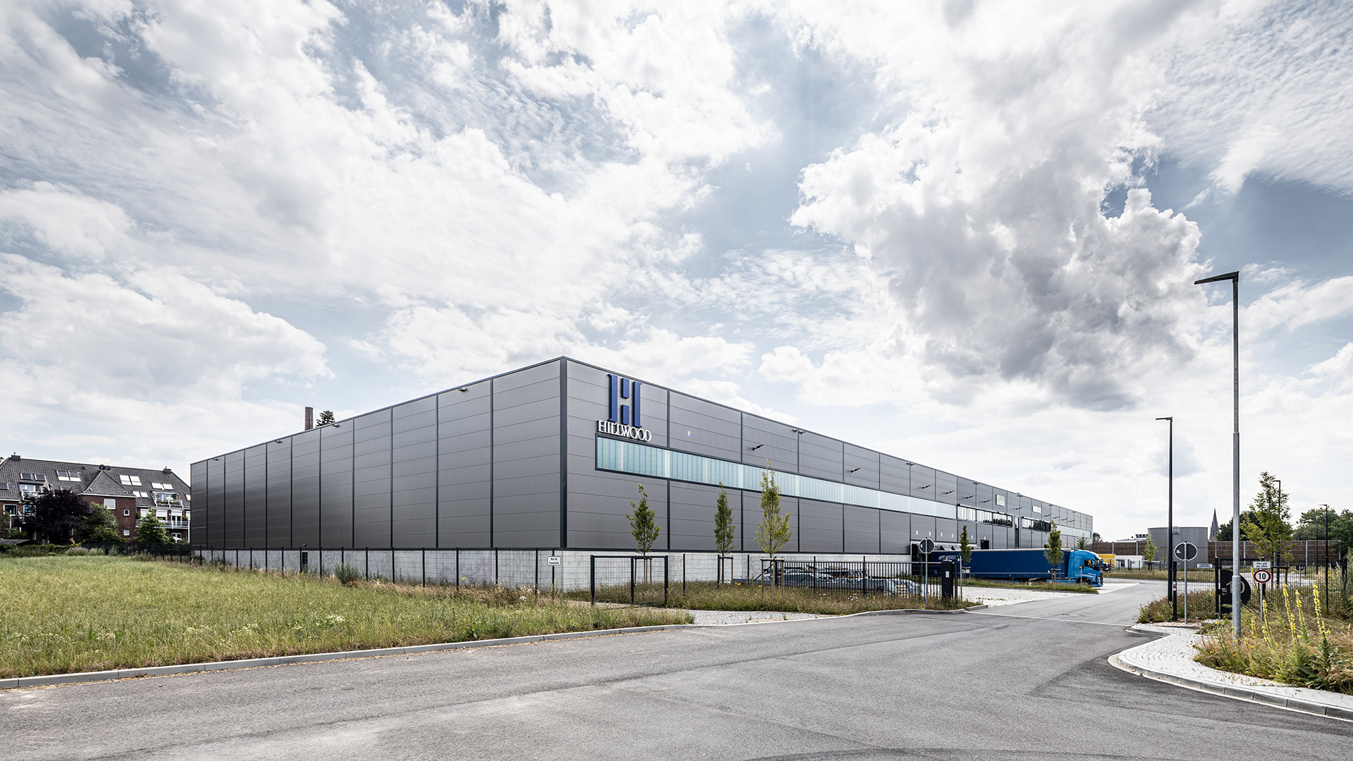 Moderne Industriehalle mit grauer Fassade und gläserner Front, umgeben von Grünflächen und Straßen. Wolken am Himmel.