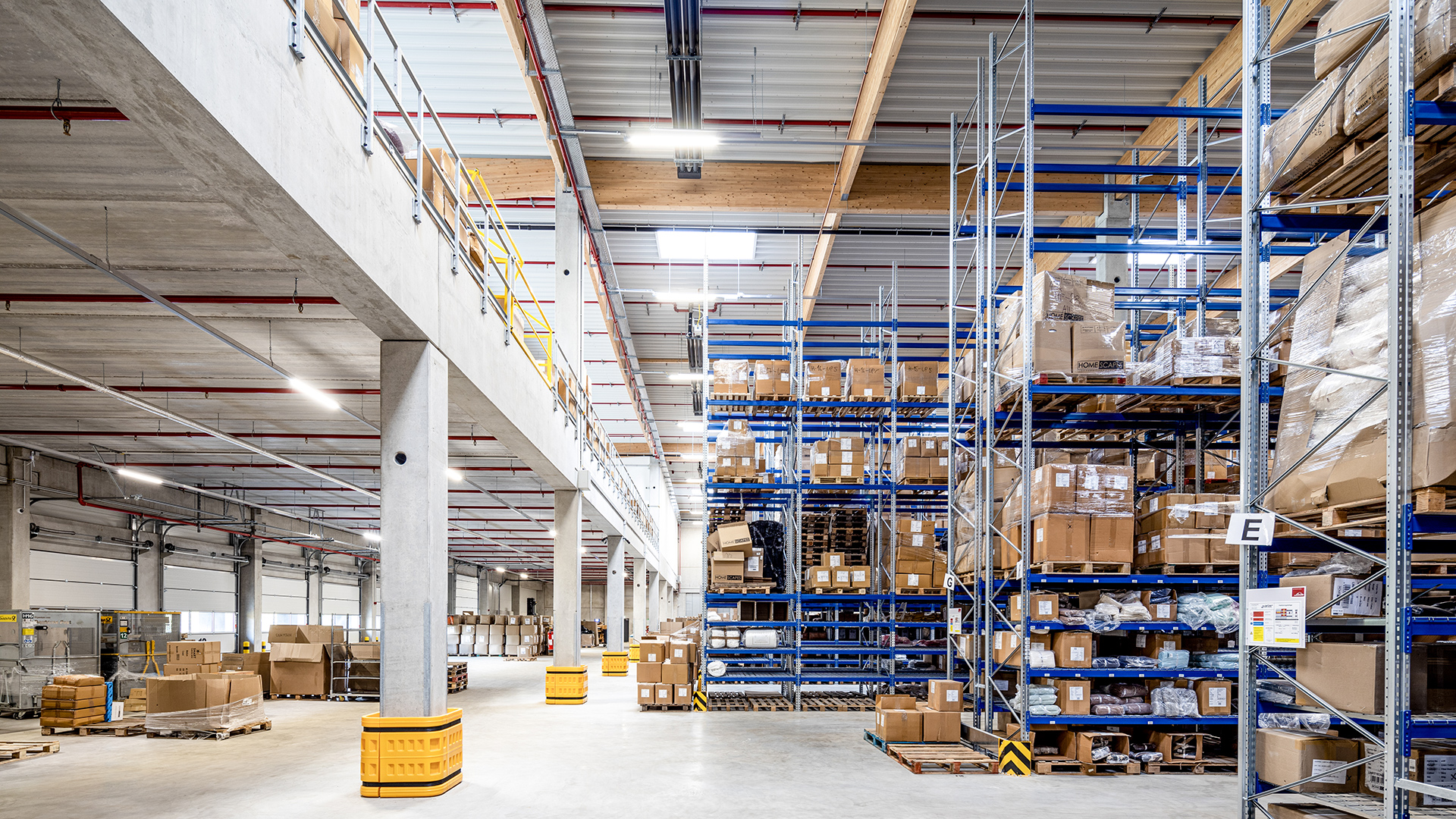Lagerhalle mit hohen Regalen, gefüllt mit Kartons, hell erleuchtet, Betonboden und Logistikmaterialien im Vordergrund.