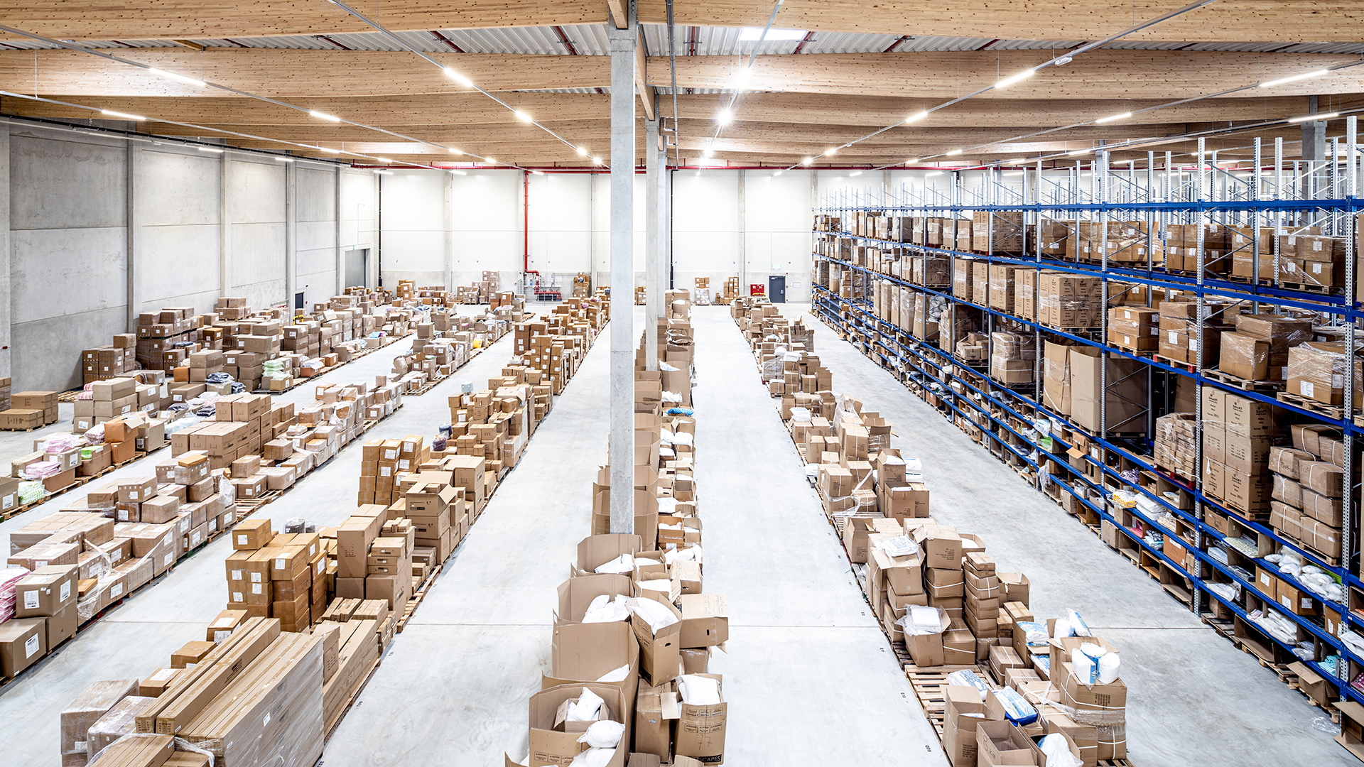 Lagerhalle mit vielen Kartons auf dem Boden und Regalen, hell beleuchtet und gut strukturiert.