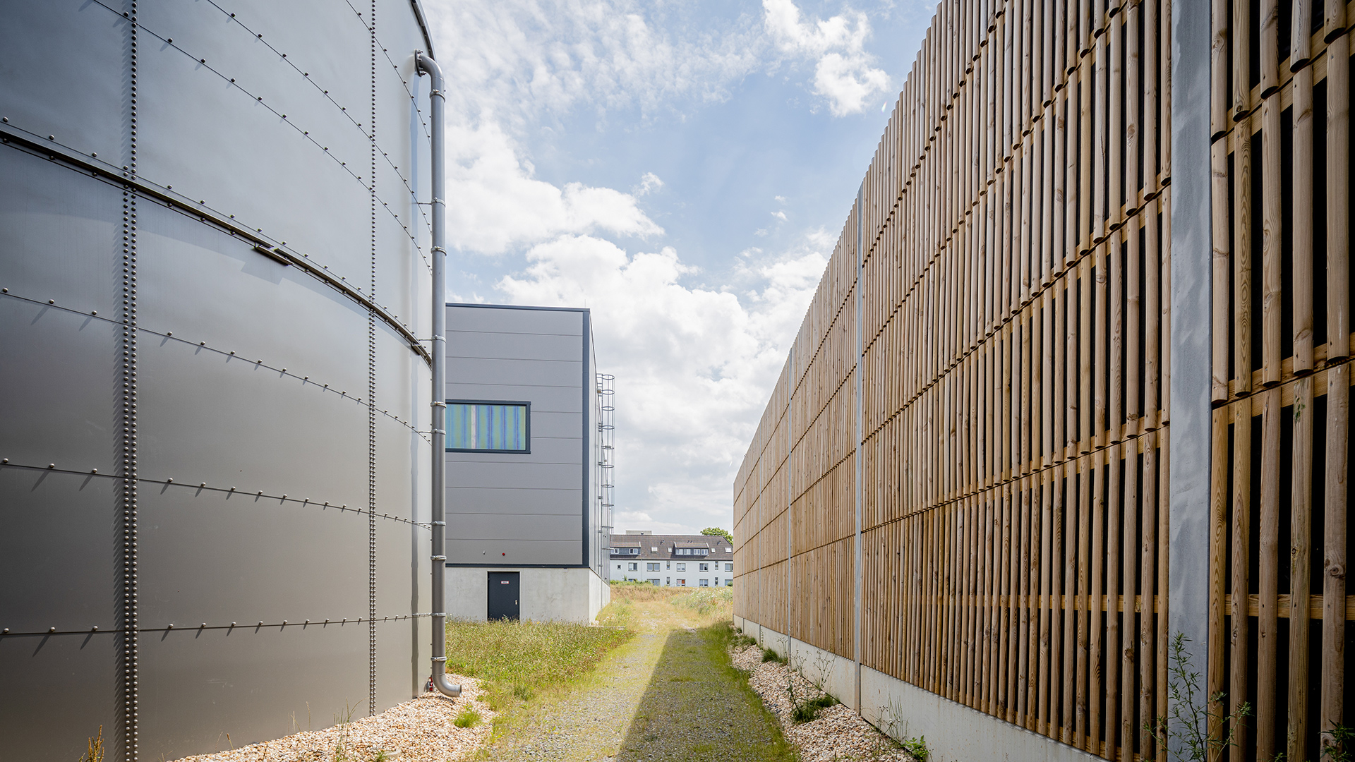 Zwei Gebäude in einem schmalen, grünen Durchgang unter blauem Himmel mit Wolken.