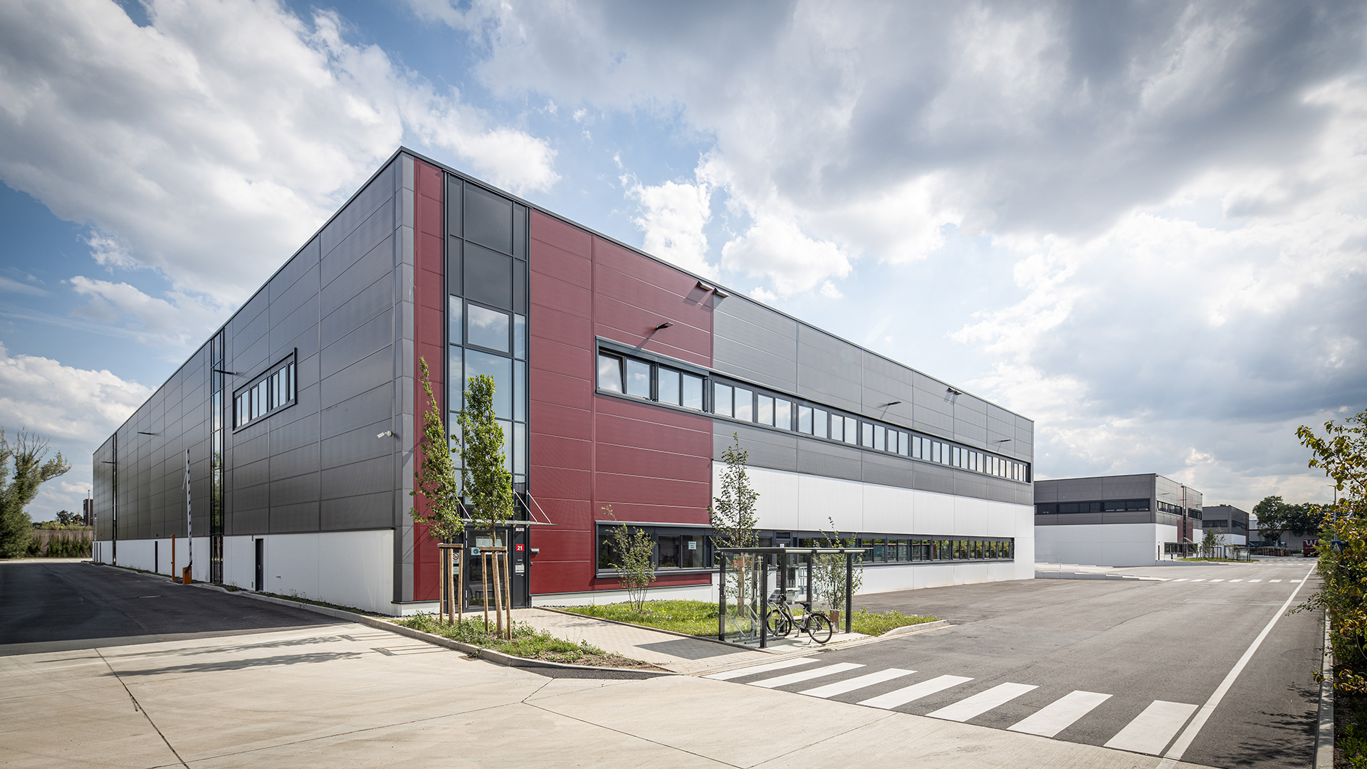 Moderne Lagerhalle mit großzügigen Fenstern und einem klaren Design, umgeben von einer Zufahrt und Stadtlandschaft.