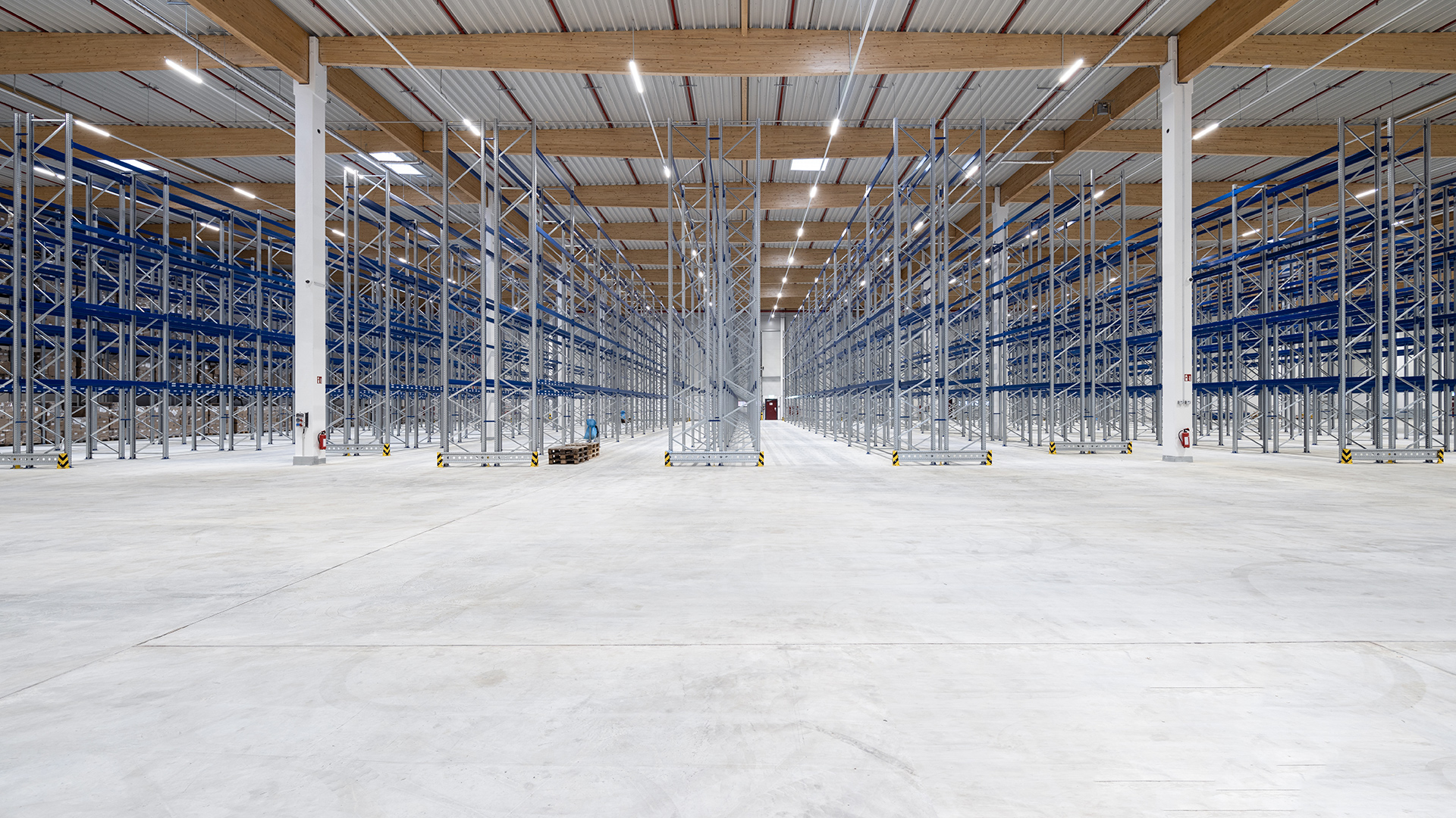 Leere Lagerhalle mit hohen Regalen auf der linken und rechten Seite, helle Beleuchtung und grauer Boden.