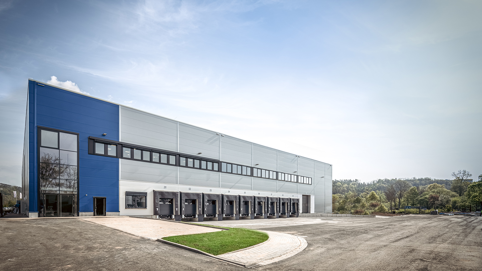 Industriegebäude mit blauer Fassade und Laderampen, umgeben von einem Parkplatz und Bäumen im Hintergrund.