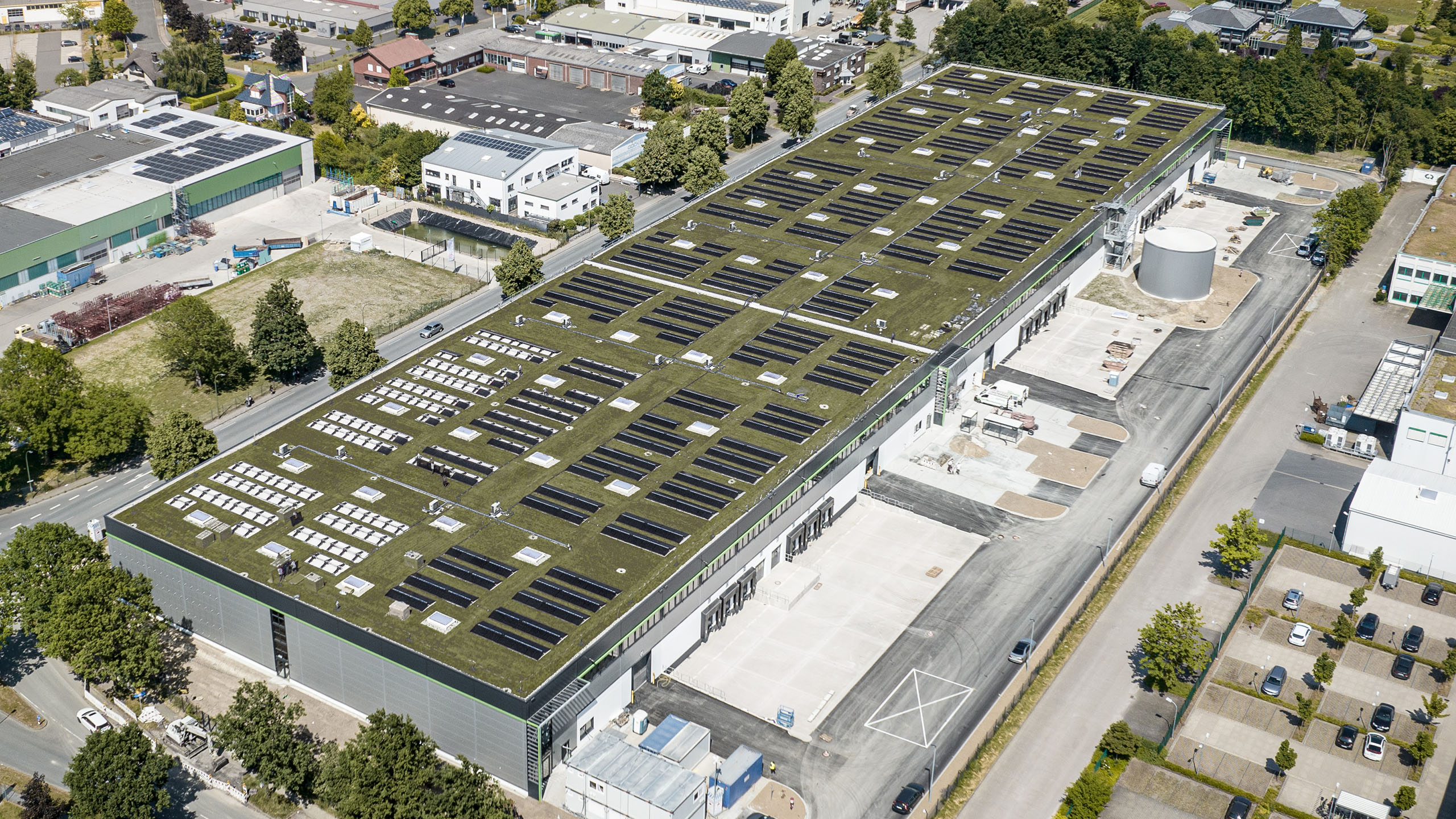 Luftaufnahme eines modernen Industriegebäudes mit grüner Dachbegrünung und Solarpanelen. Umgebung mit Bäumen und Straßen.