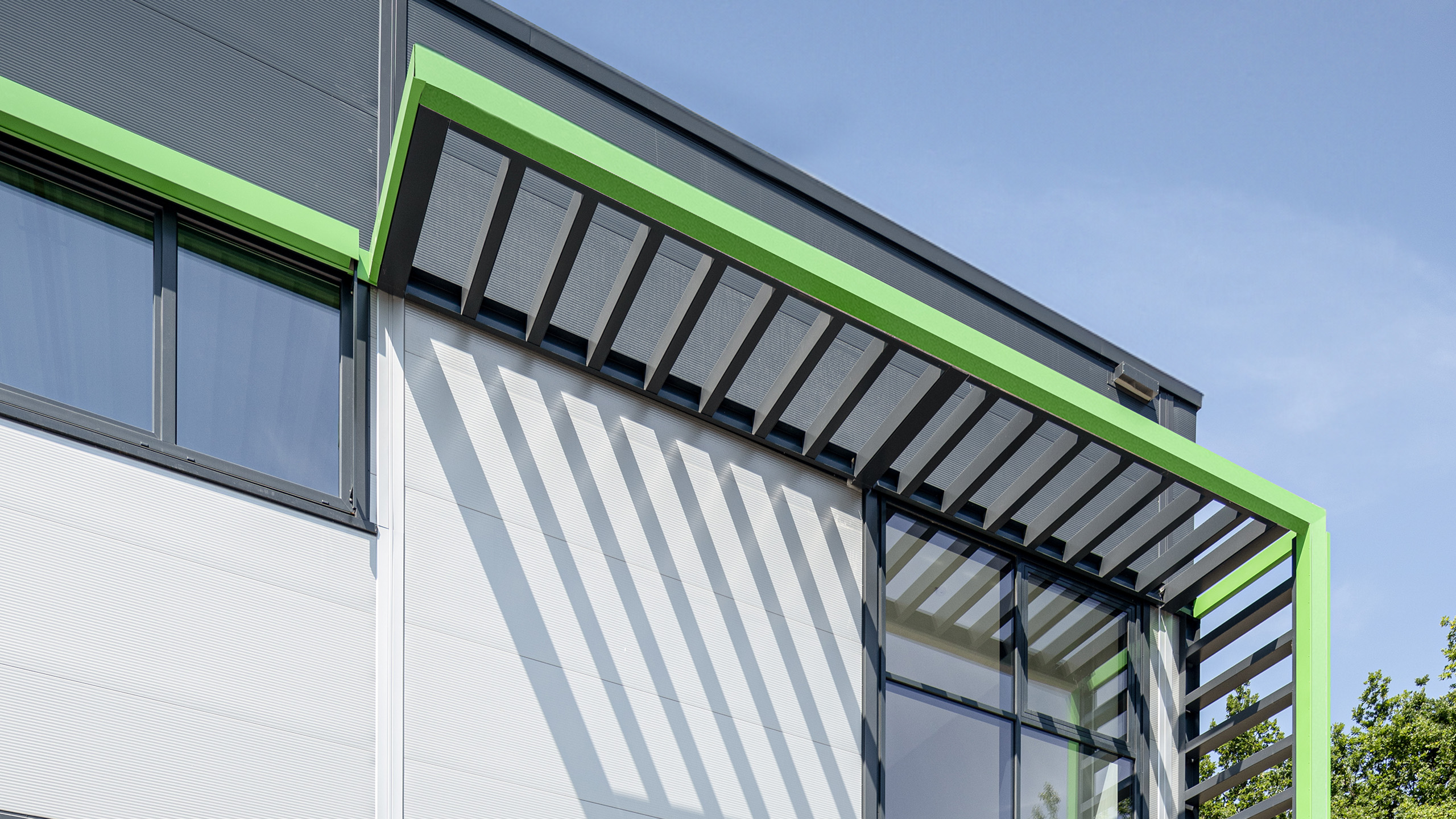 Moderne Gebäudeansicht mit grünem Dachvorsprung, großen Fenstern und Schattenwurf unter blauem Himmel.