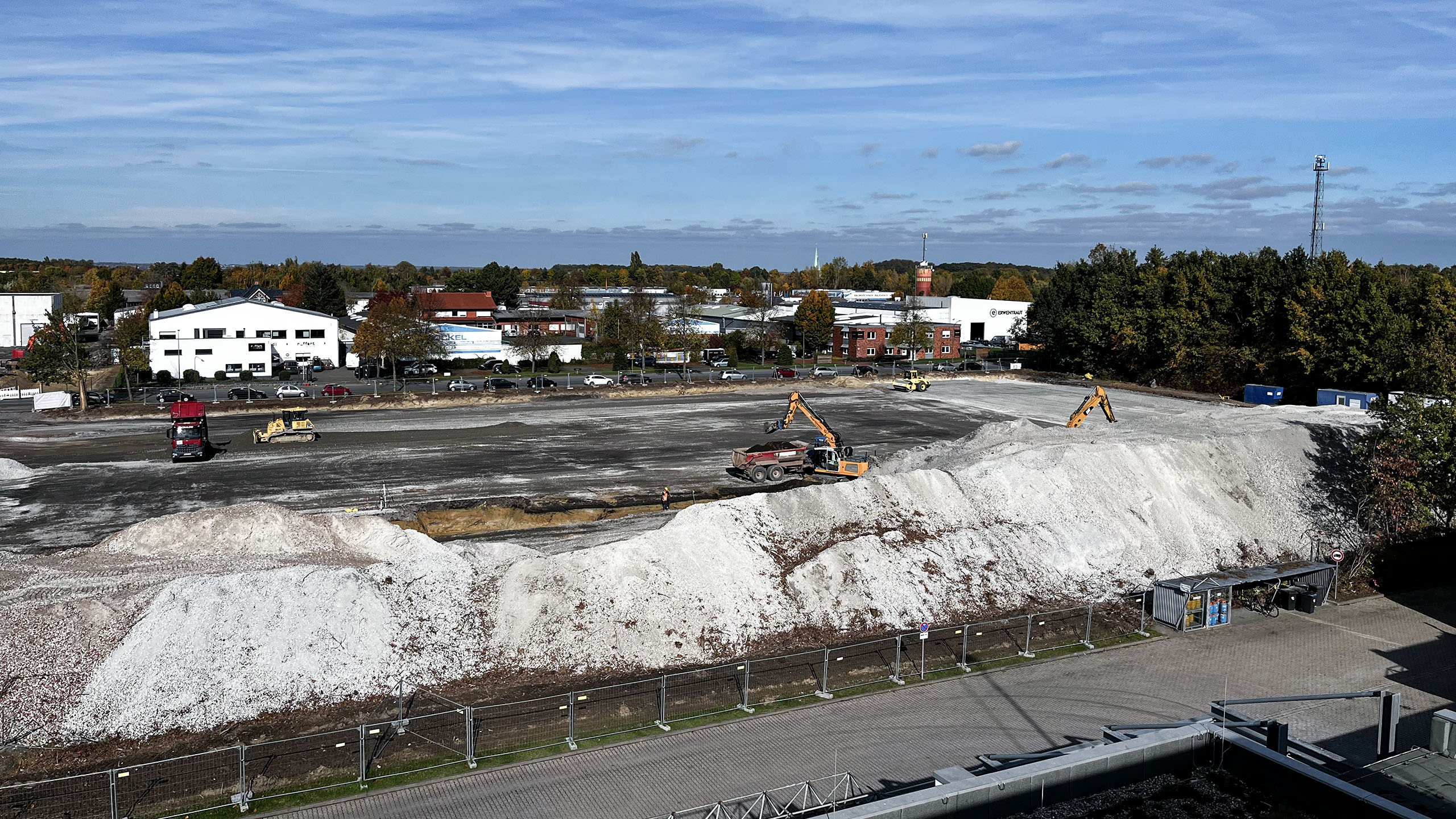 Bauplatz mit Erdbauten, Baggern und Lkw, umgeben von Gewerbegebäuden und Bäumen, unter blauem Himmel.