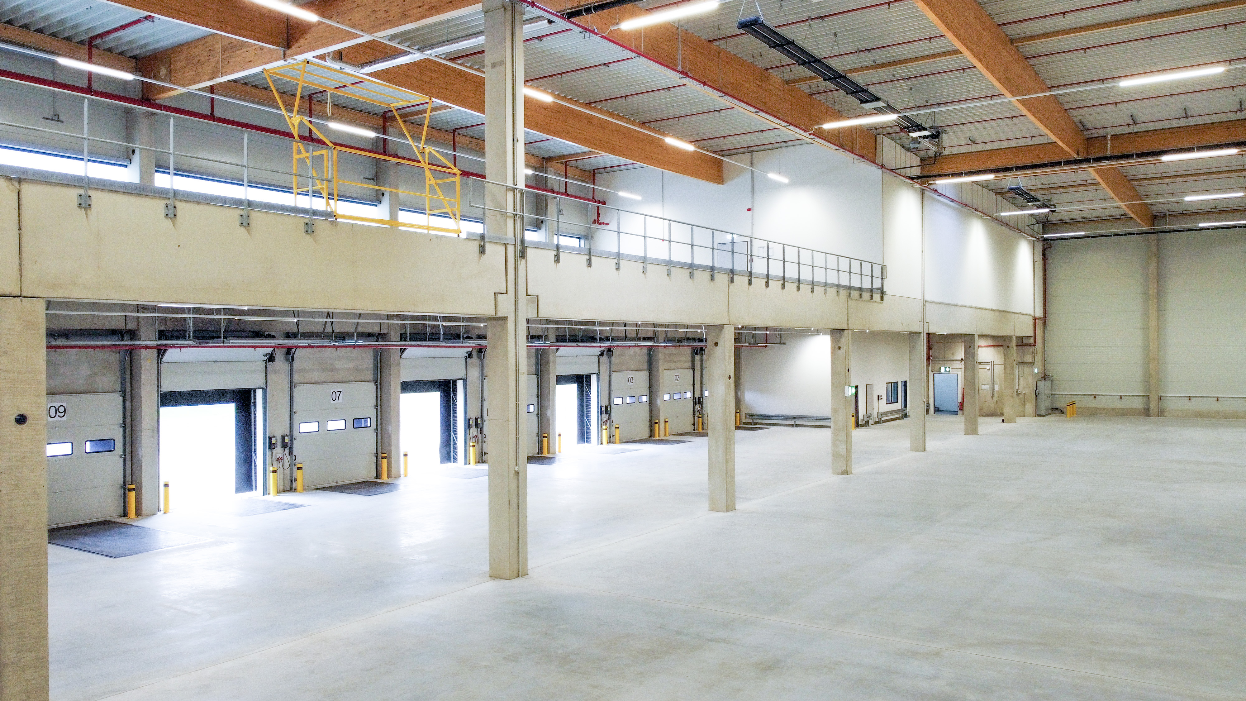 Geräumige Lagerhalle mit Rolltoren, hohen Decken und neutralem Bodenbelag. Besondere Merkmale sind Stahlträger und eine Galerie.