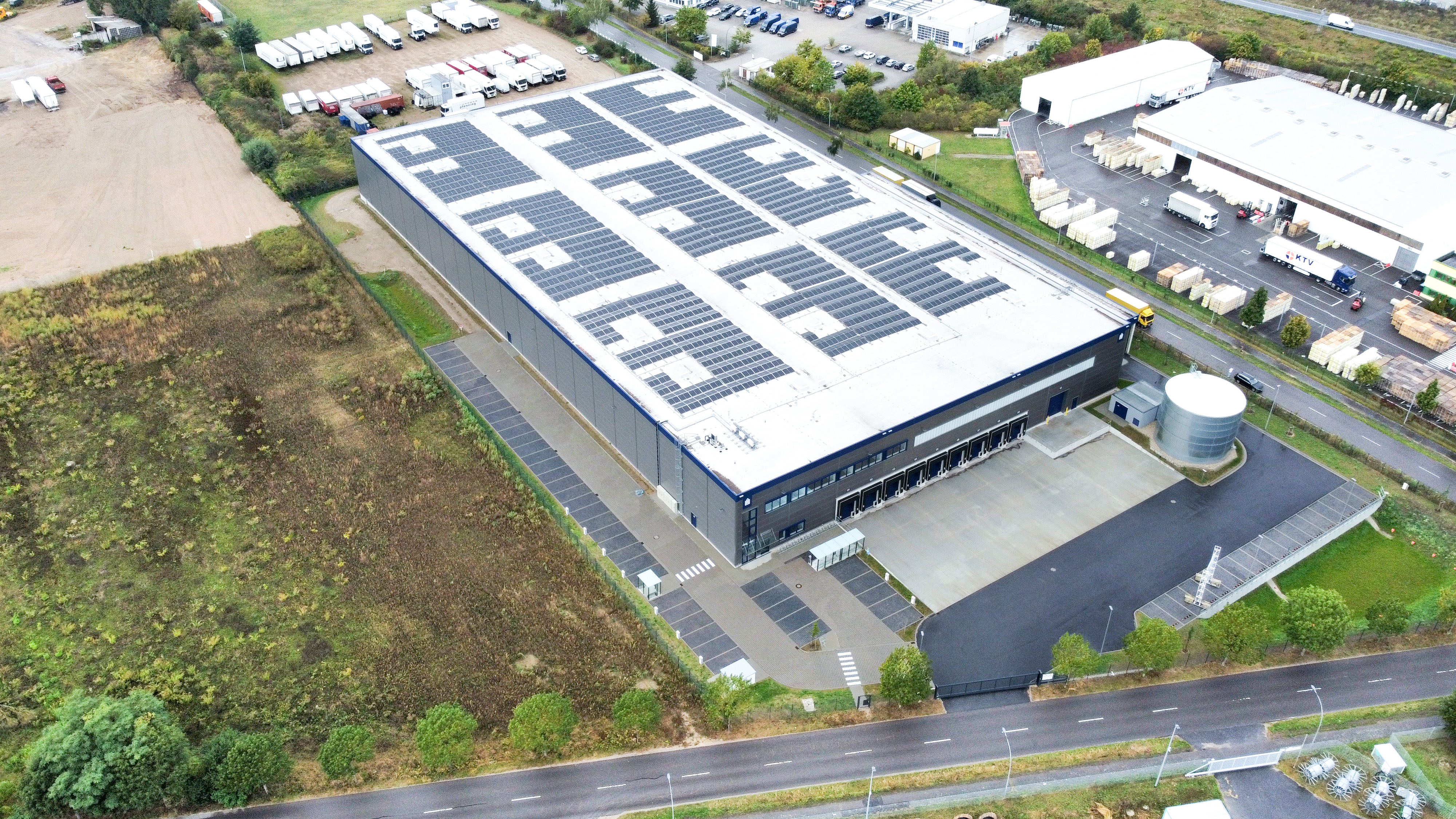 Dachansicht eines modernen Lagerhauses mit Solarpanelen und Parkplatz, umgeben von Bäumen und Industriegebäuden.