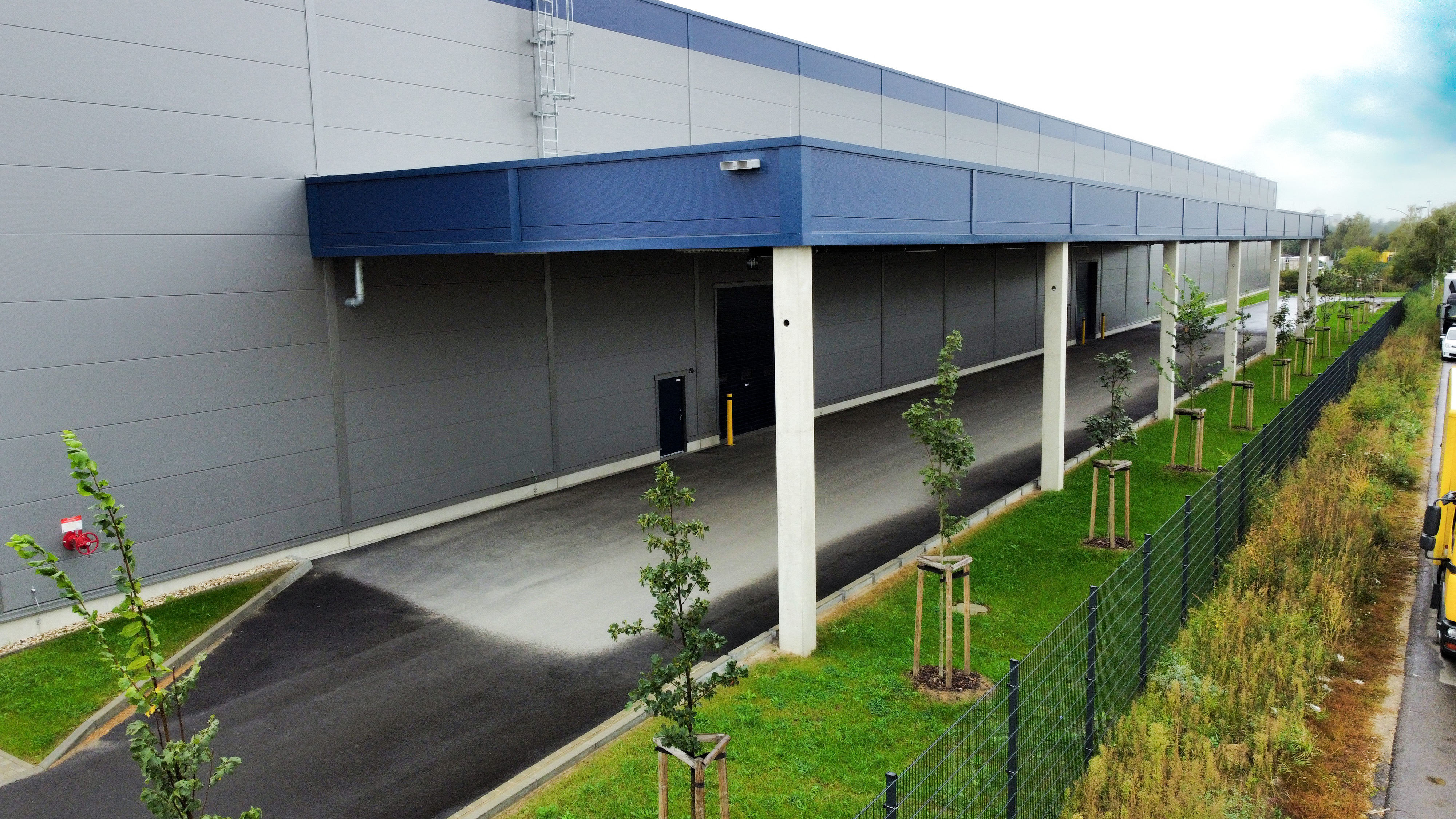 Lagergebäude mit überdachtem Eingang, Asphaltweg, neben Bäumen und Zaun auf einer grünen Fläche.