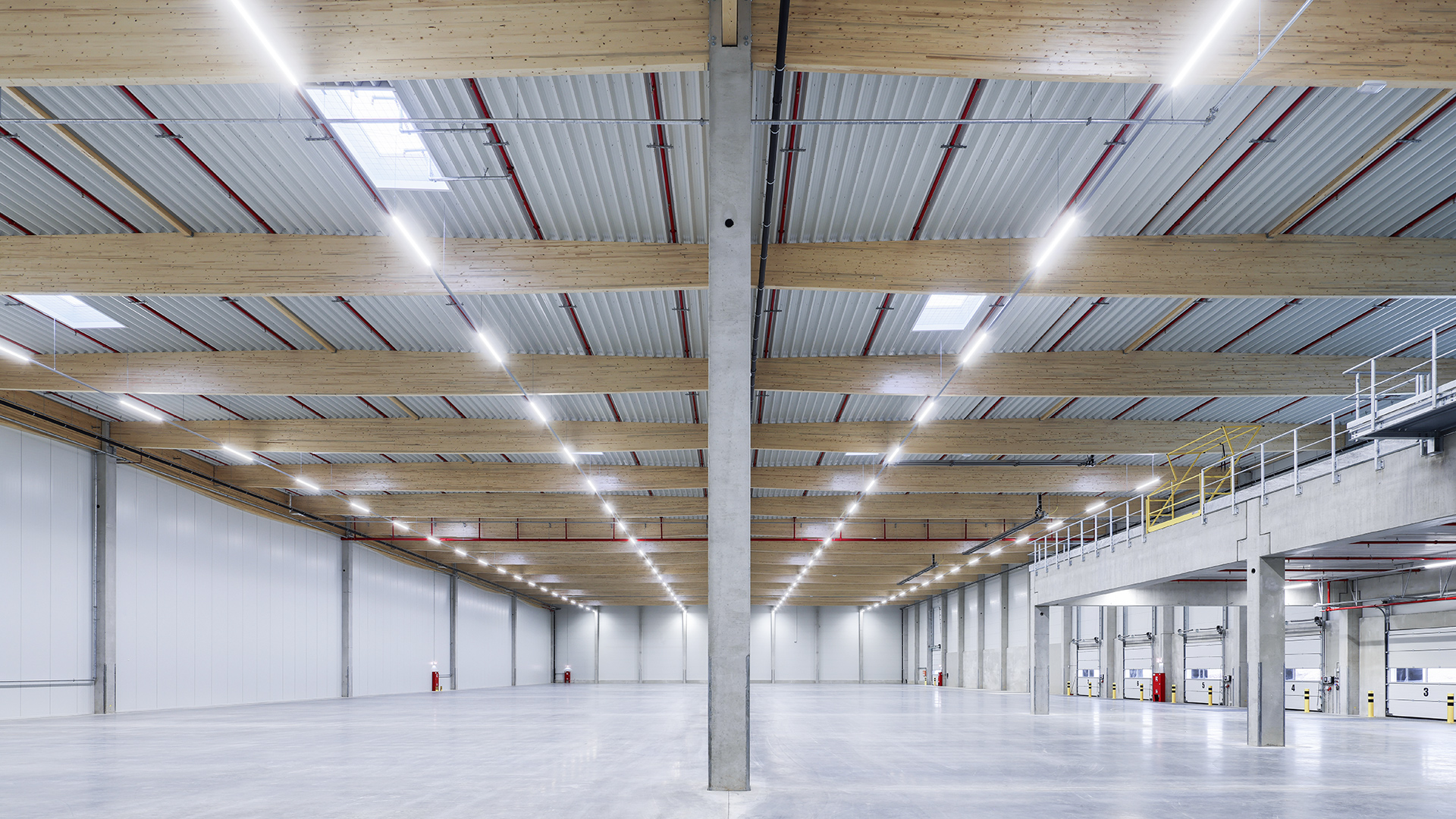 Weißer Lagerraum mit Holzbalken und betongegossenen Säulen, beleuchtet von Deckenleuchten.