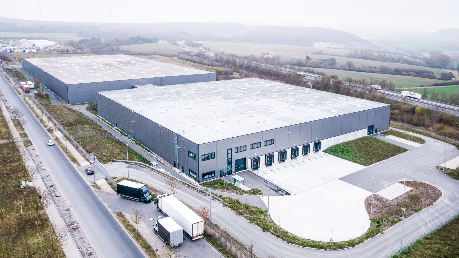Luftbild eines modernen Logistikgebäudes mit Laderampen, umgeben von einer grünen Landschaft und einer Straßenanbindung.