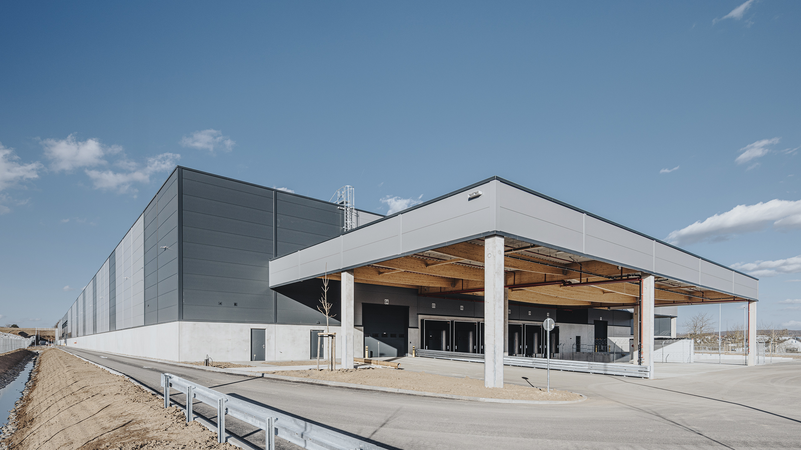 Großes modernes Lagergebäude mit Vordach, grauer Fassade und klarem blauen Himmel im Hintergrund.
