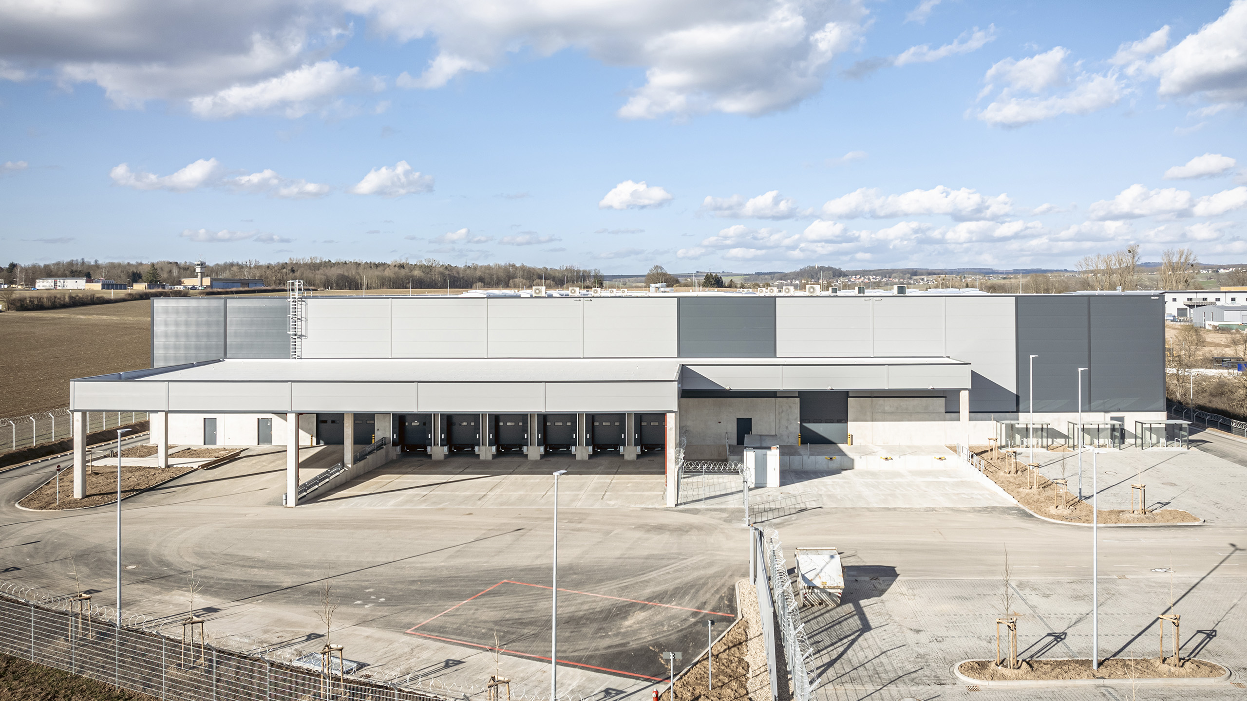 Großes modernes Lagergebäude mit mehreren Auffahrten und Parkplatz, umgeben von weitem Freiraum und sanftem Himmel.