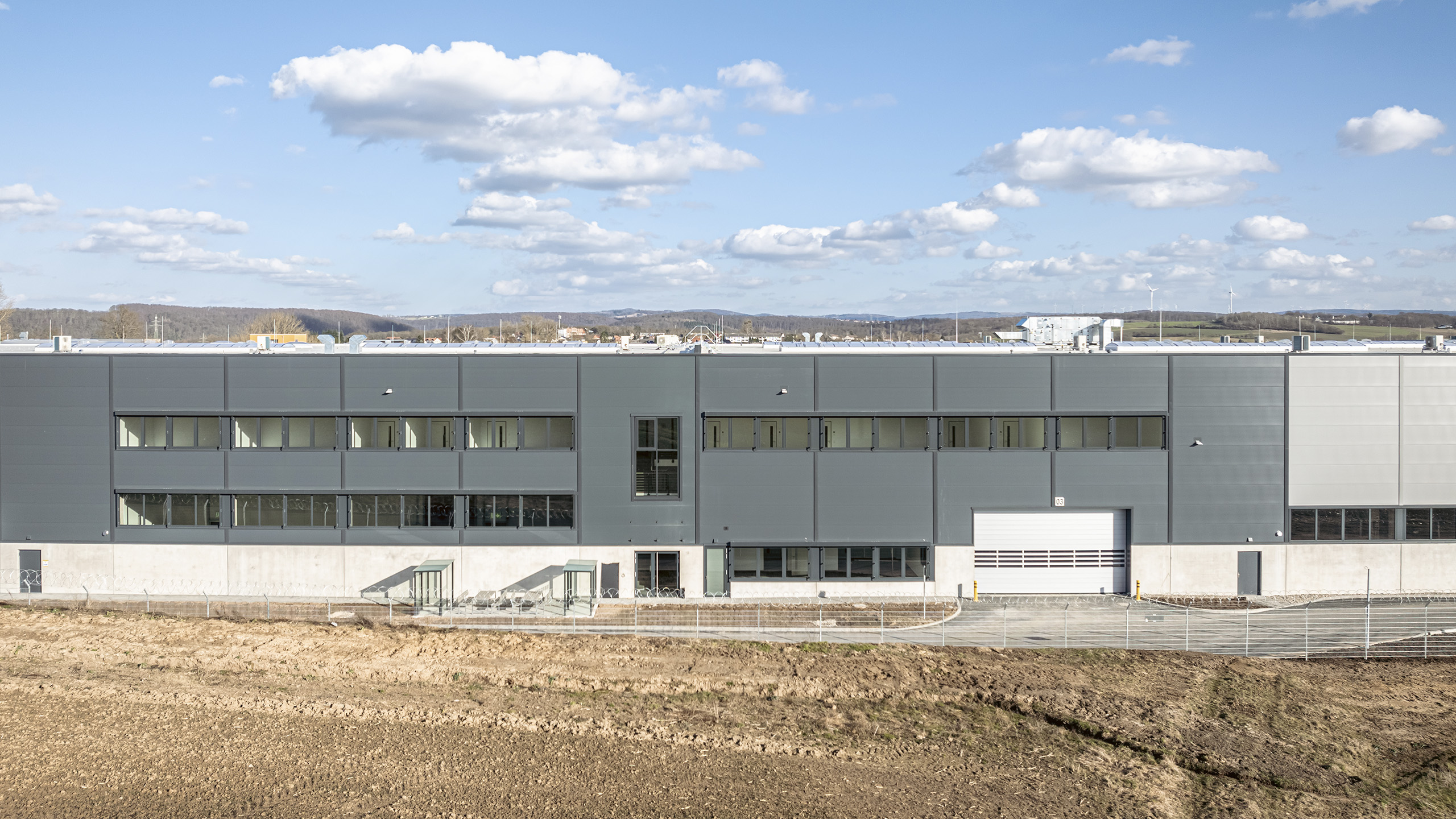 Moderne gewerbliche Gebäude mit grauer Fassade, umgeben von freiem Gelände und blauen Himmel, mit Wolken im Hintergrund.