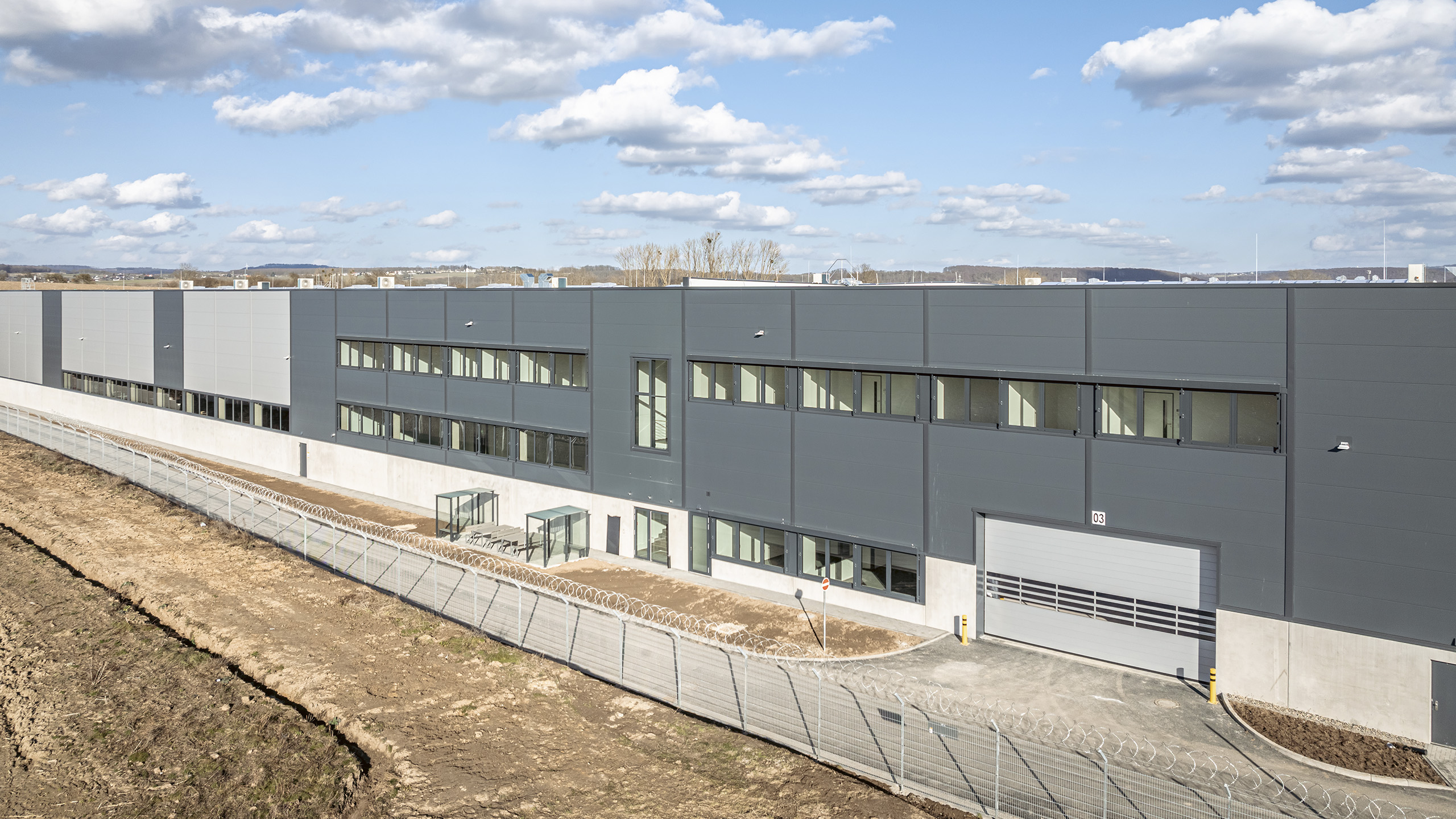 Moderne Industriegebäude mit großen Fenstern, umgeben von einem Zaun, unter blauem Himmel mit weißen Wolken.