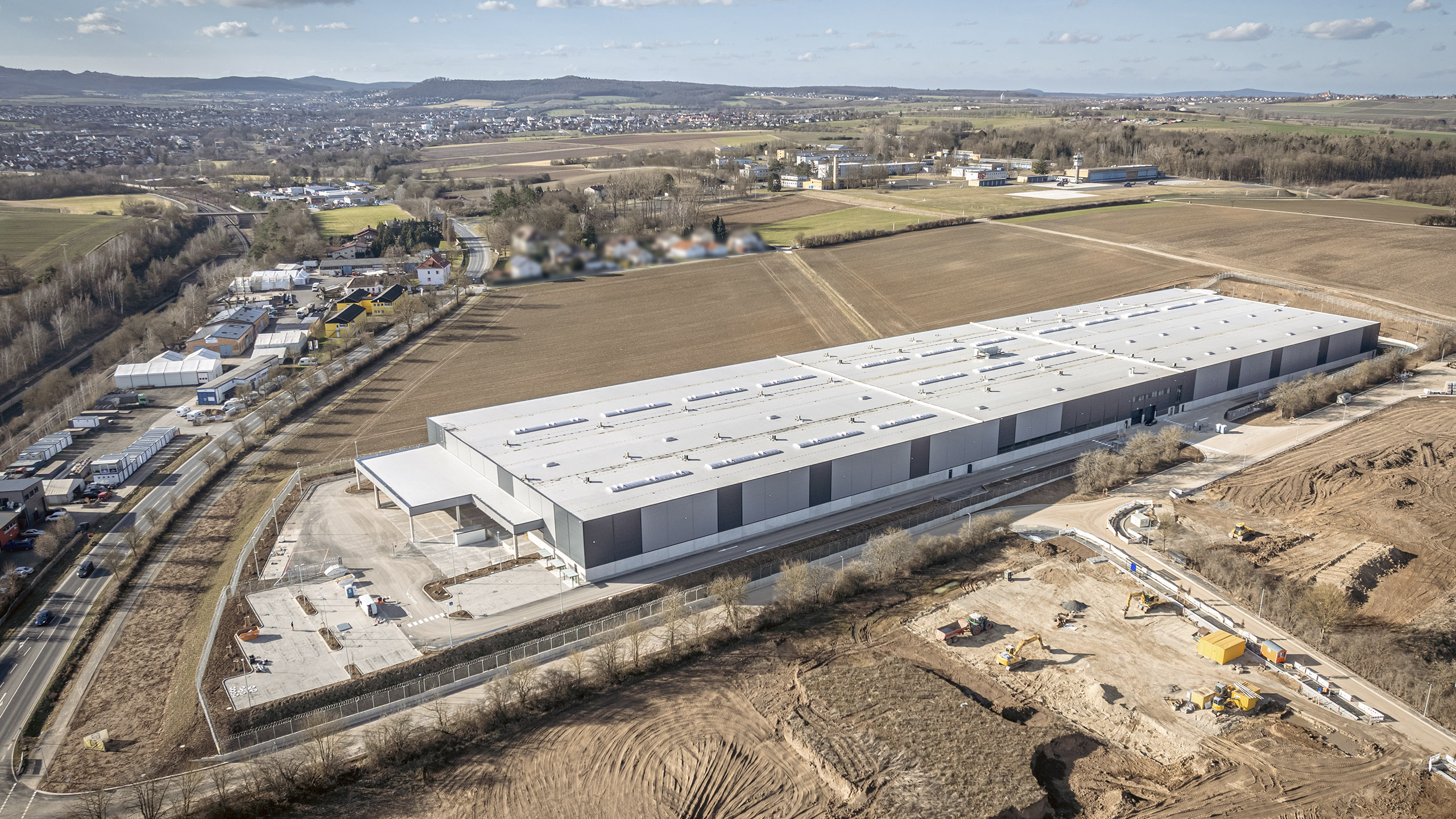 Luftaufnahme eines modernen Logistikgebäudes auf einem großen Grundstück, umgeben von Feldern und Stadtansichten.