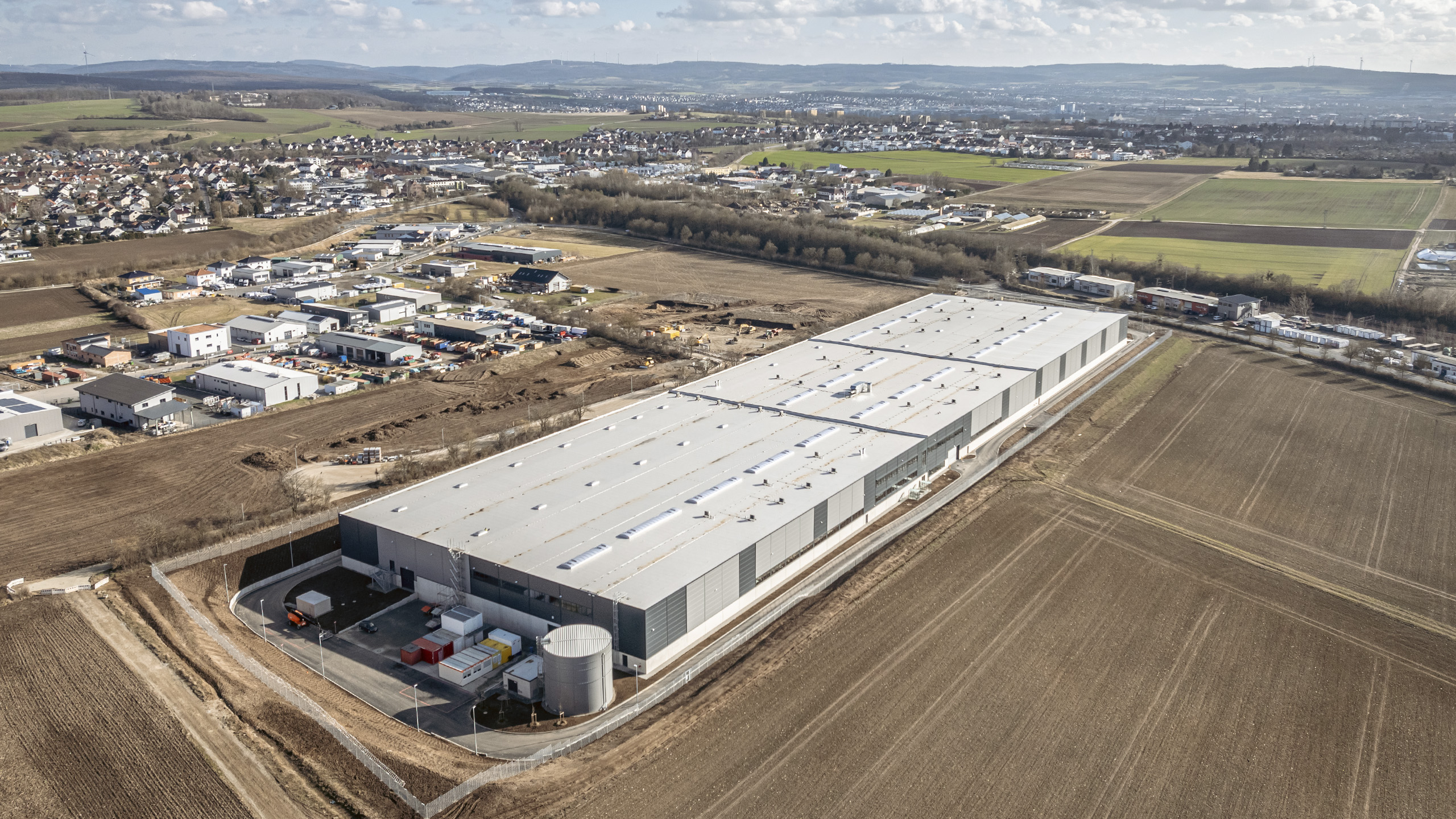 Luftaufnahme eines großen Lagergebäudes umgeben von Feldern und vereinzelten Wohnhäusern.