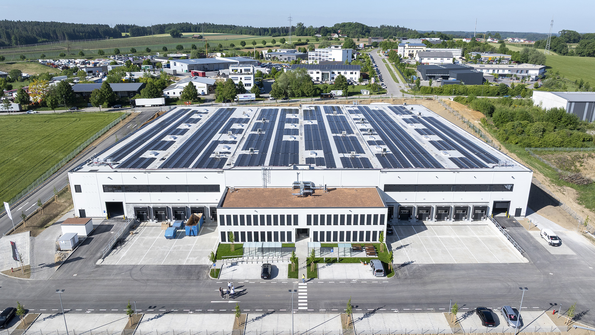 Luftaufnahme eines modernen Gebäudes mit Solarpanels, umgeben von Grünflächen und Parkplatz.