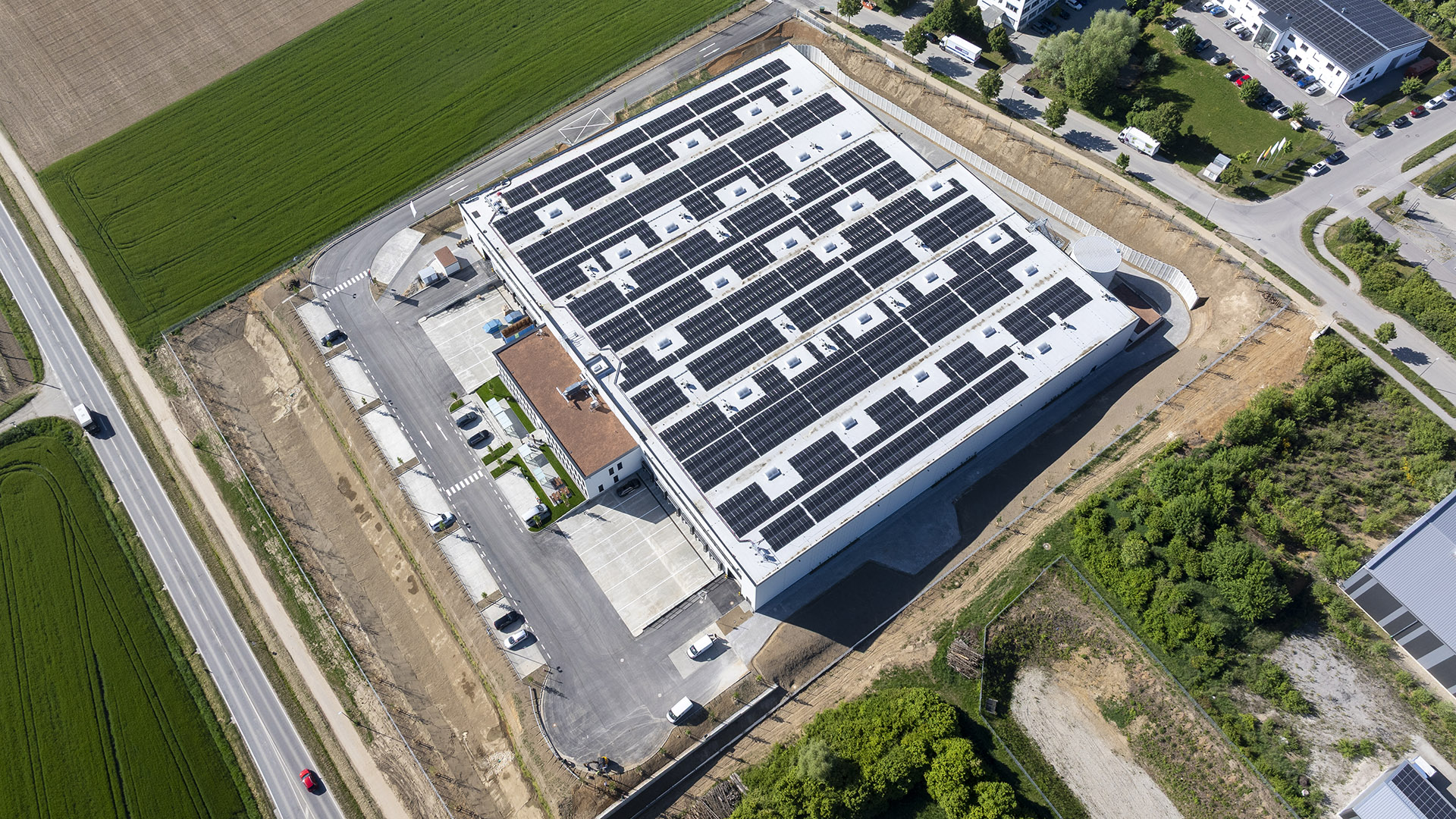 Dachansicht eines modernen Gebäudes mit Solarpanelen, umgeben von Straßen und Feldern.
