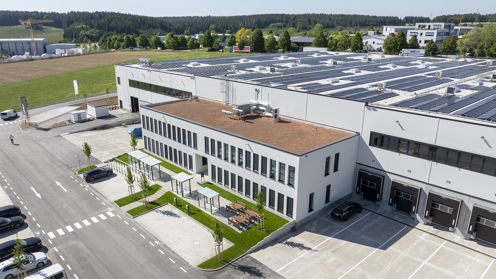 Moderne, flache Unternehmensgebäude mit begrünten Flächen, Parkplätzen und Solarplatten auf dem Dach.